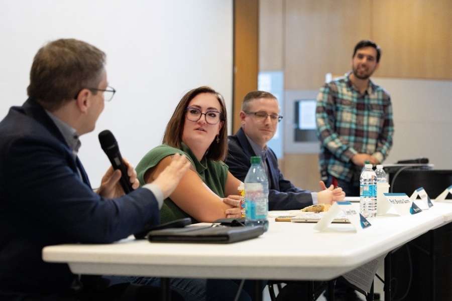 Panel discussion with three speakers seated at a table and one standing behind, as a man speaks into a microphone during an indoor event.