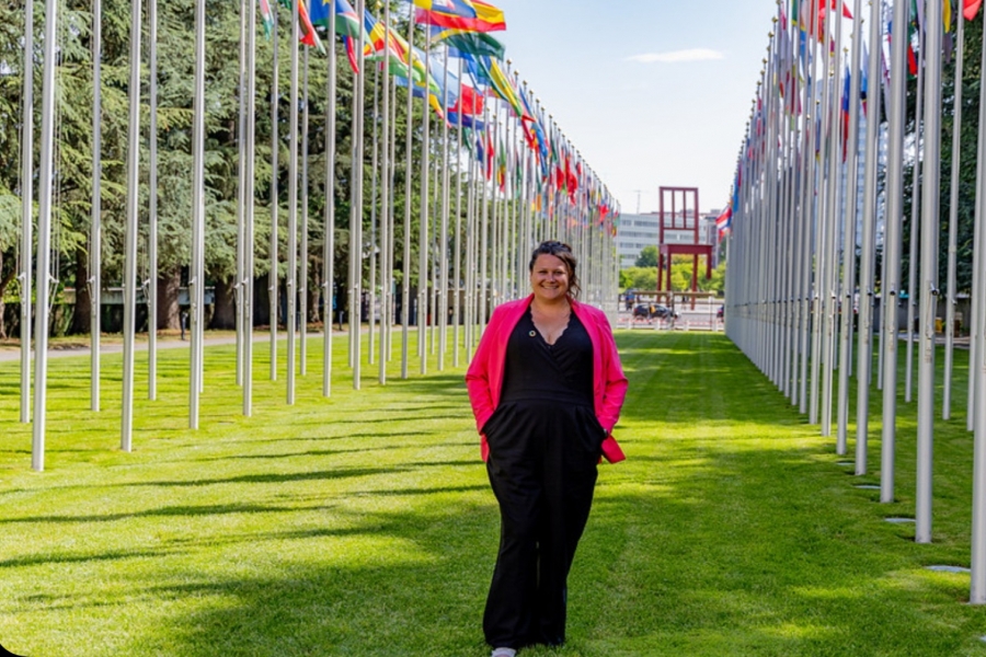 A woman standing in a grassy area with two rows of large white flagpoles with flags of various countries in a line on either side of her.