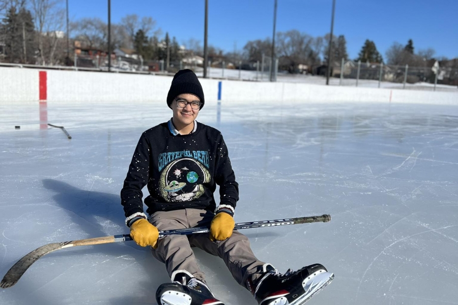 male sits on ice in outdoor rink on a sunny day with hockey stick in hands, wearing ice skates.