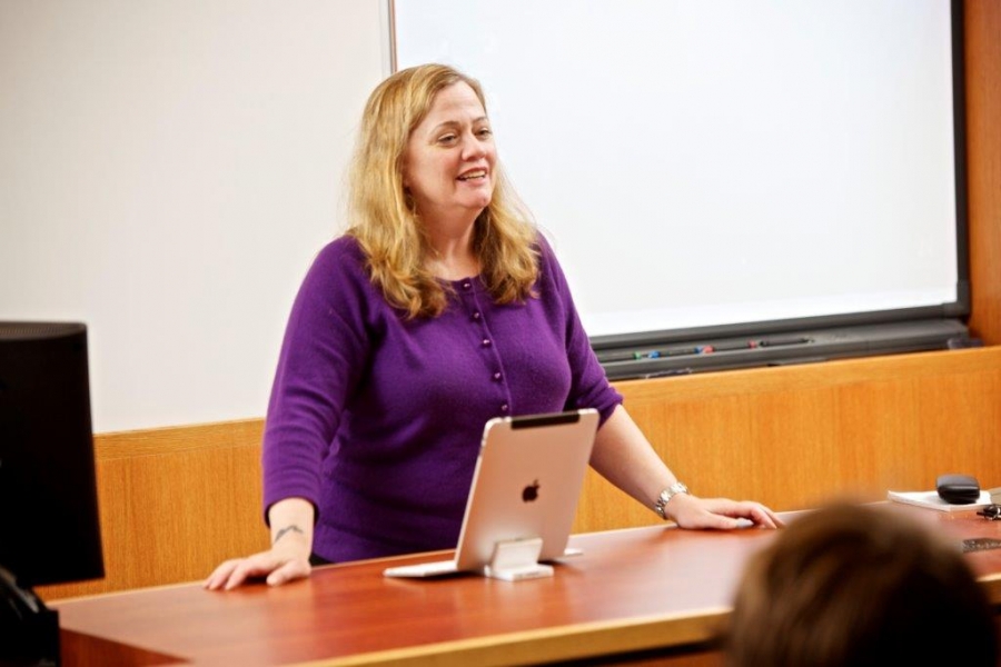 A woman in a purple sweater stands behind a classroom desk, in action teaching a class