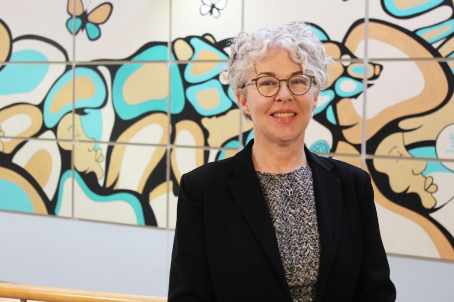 Kellie Thiessen stands in front of a colourful Indigenous mural at the College of Nursing.