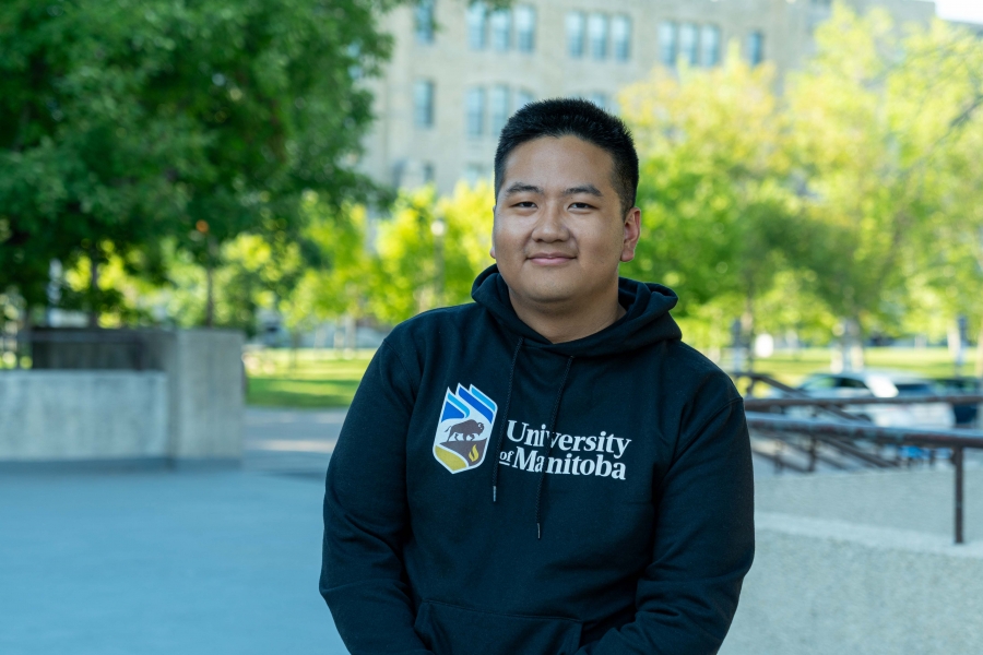 A student wearing a University of Manitoba hoodie posing in front of a background of university buildings and green trees.