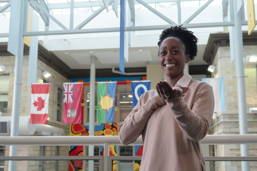 A person strikes the Give to Gain pose: holds one hand to the heart, the other hand reaching out. In the background are flags suspended from the ceiling.