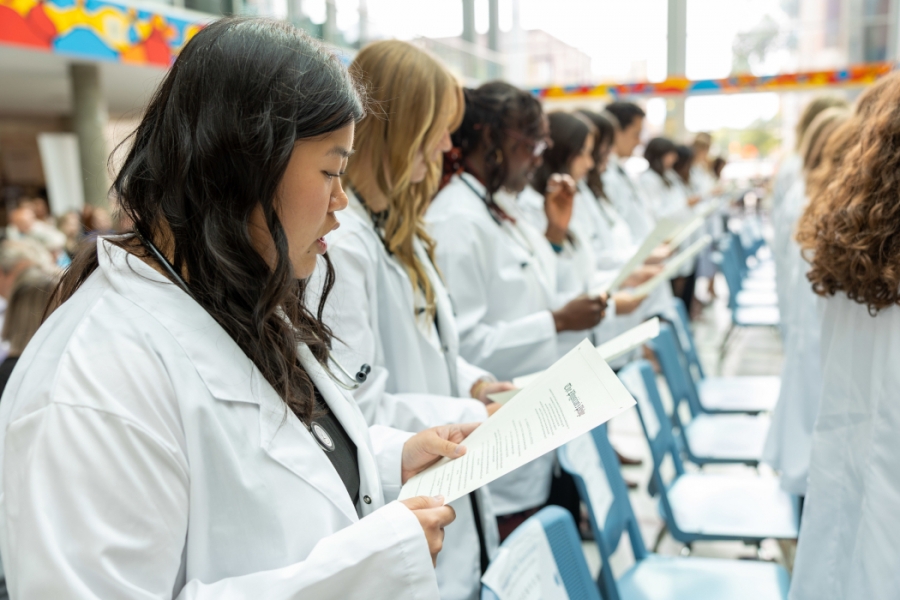 Students wearing white coats and stethoscopes around their necks, read off of sheets of paper.