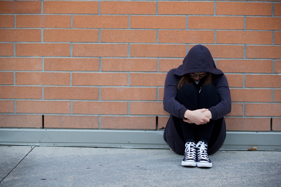 A youth sits against a wall with a hood covering their face. 