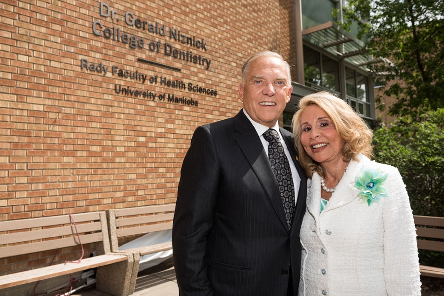 Two people stand in front of the dentistry building. A sign on the building reads "Dr. Gerald Niznick College of Dentistry. Rady Faculty of Health Sciences. University of Manitoba."