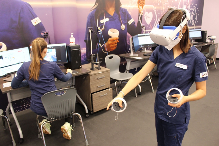 A nursing student wears a virtual reality headset and hand controls while another student works at a computer station behind her.