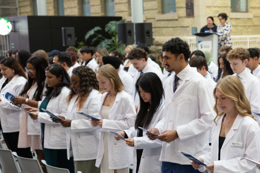 Mary Bertone and Dr. Anastasia Kelekis-Cholakis stand on a stage at a lectern in the background. Rows of students wearing white coats read off of papers.