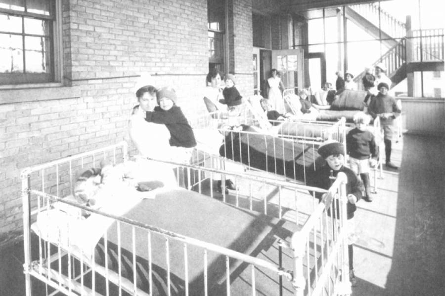 Black and white photo of an old hospital. Kids sleep in metal cribs and nurses hold children.