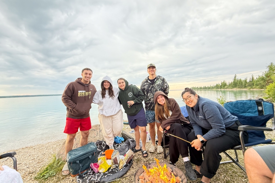 A group of six people gathered around a campfire beside a lake.