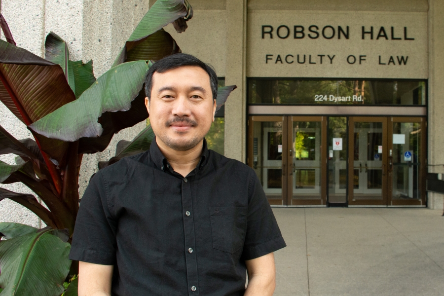 Rolan Tan wears a black shirt and smiles in front of a green leafy plant in front of Robson Hall