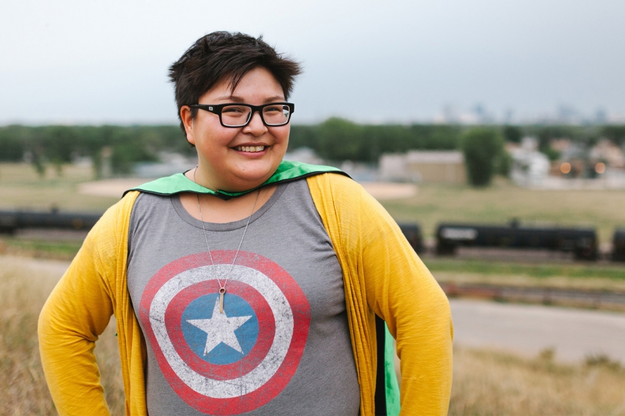 Woman wearing glasses, a tshirt with the Captain America shield on it and a cape, stands with her hands on her hips.