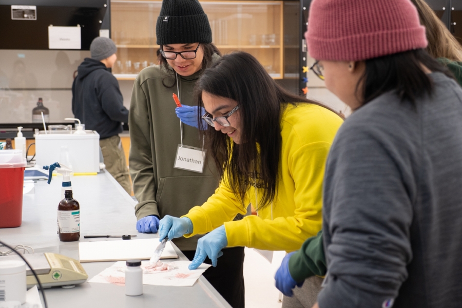 One student mixes ingredients on a table in a lab, while three other students watch.