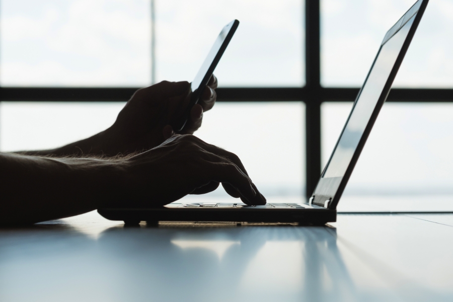 Backlit photo of someone using a phone and a laptop.