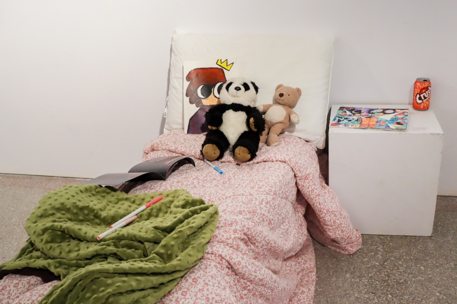 A bedinstallation with stuffed animals, blankets, drawings, and sketchbooks arranged as part of the exhibition.