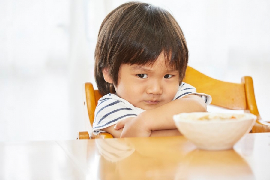 A child that looks unhappy sits at a table with their arms crossed. A bowl of food sits on the table.