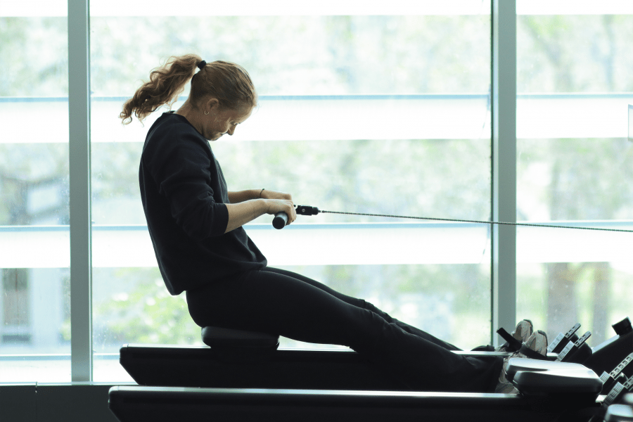  A young women works out on a rowing machine with big windows in the background.