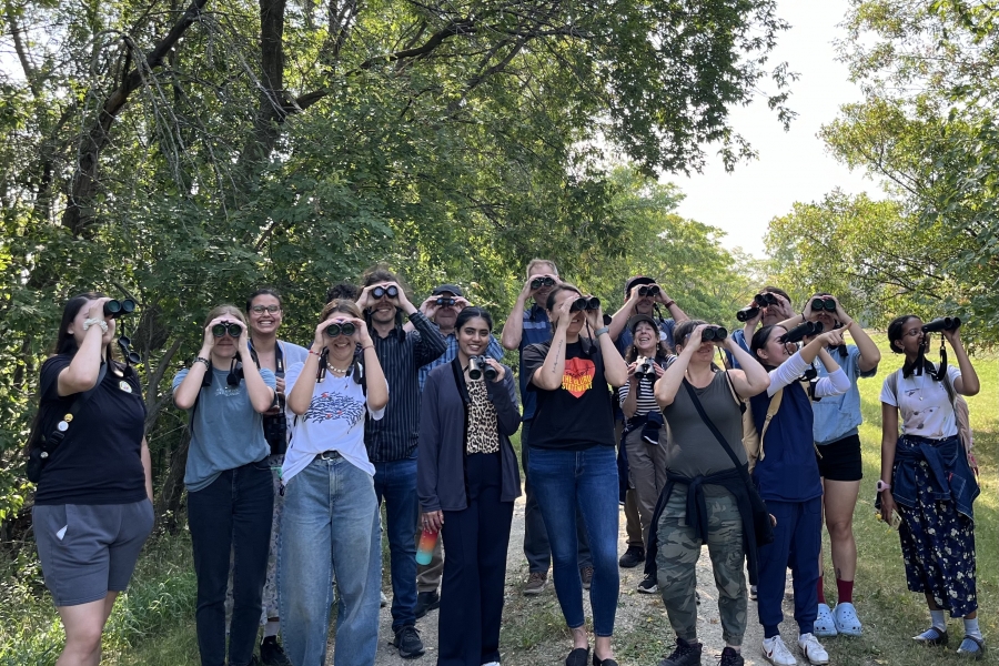 group of people holding up binoculars to their eyes and smiling, with trees in the background.