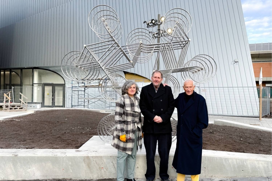 Three people stand in front of a bicycle sculpture made of 18 bicycles. The Desautels Concert Hall is in the background.