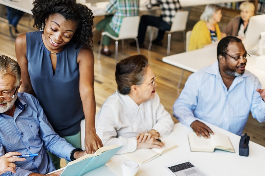 A group of diverse elderly individuals engaged in a discussion around a table with a laptop, notebooks, and documents in a library.