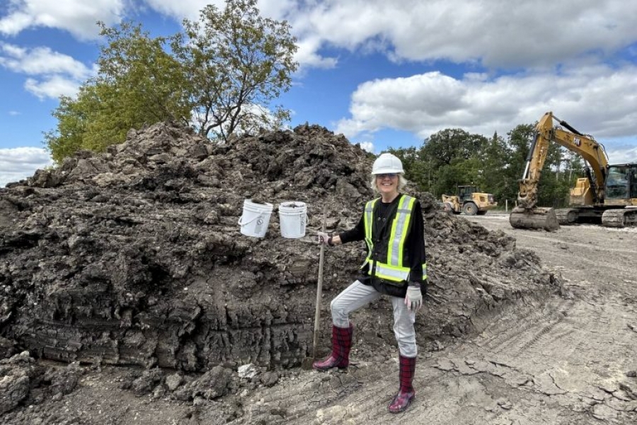 Instructor Grace Nickel stands at a construction site holding two buckets of locally sourced clay, wearing safety gear.