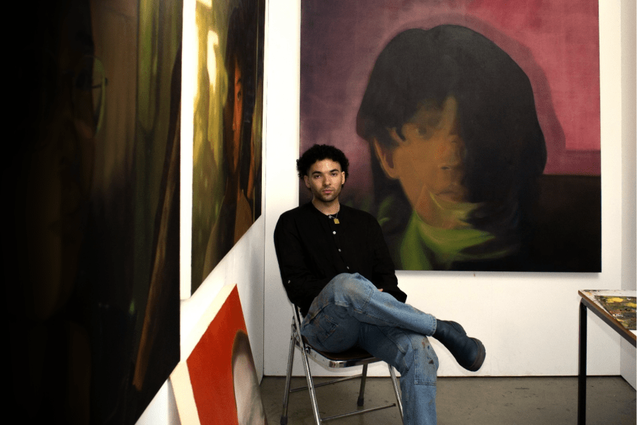 Joel Nichols sits in a studio surrounded by large-scale portrait paintings, with a paint-covered worktable nearby.