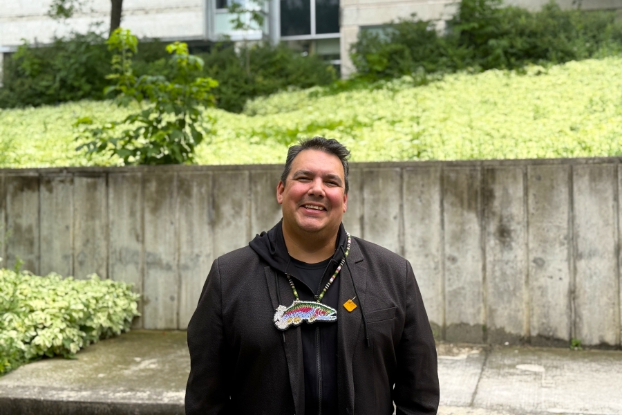 man smiles at camera, against greenery in background, wearing black sweater and beaded medallion. 
