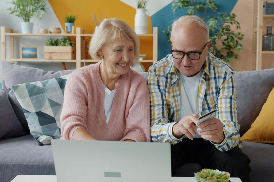 Two older adults are seated on a couch ordering an item online on their laptop.