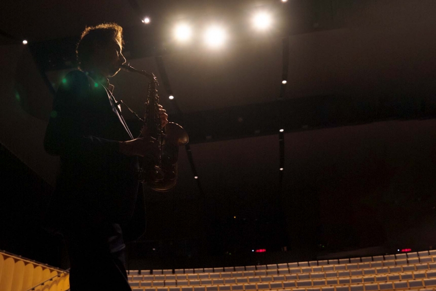 A man in a suit plays a saxophone on a dark stage.
