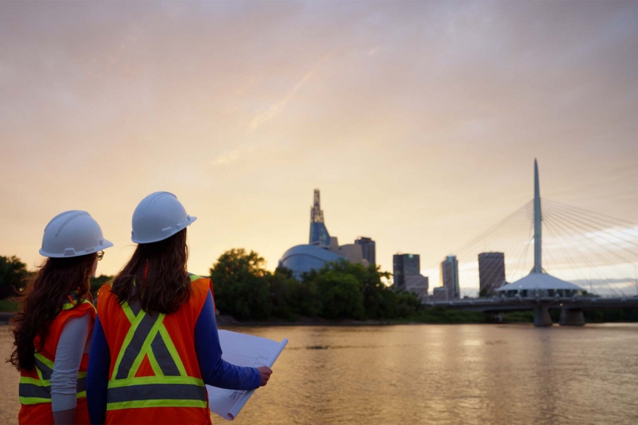 Two female engineers in safety gear look across the Red River in Winnipeg to the city's skyline.