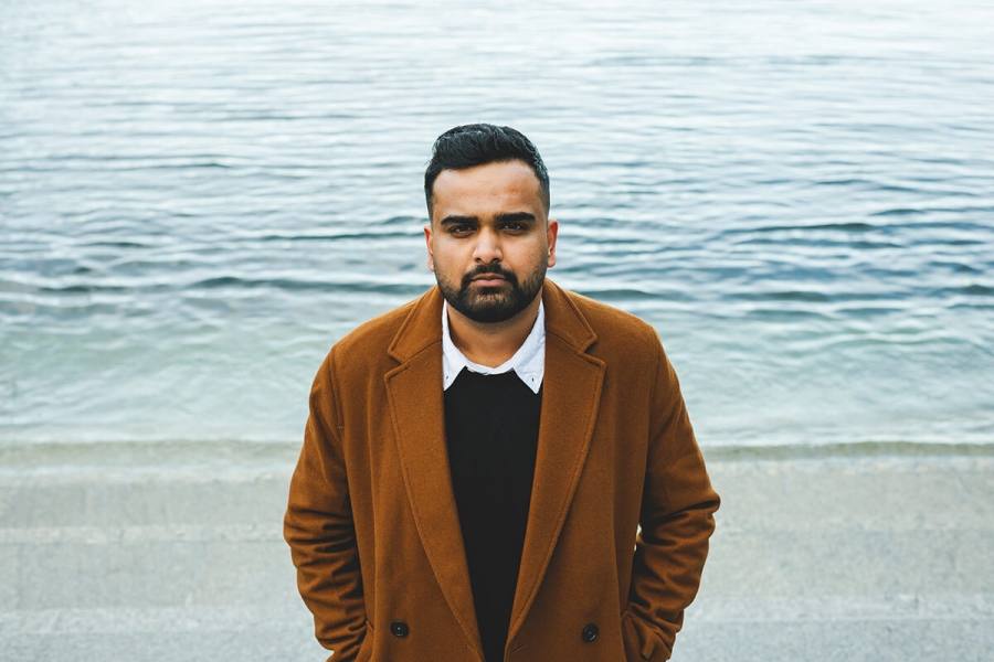 Nabil Iqbal stands in front of a body of water, his hands in his pockets, looking at the camera.