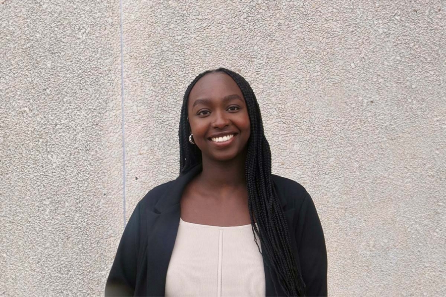 Witta Irumva, Black female student standing on front of a grey wall, wearing a black and white outfit, smiling.