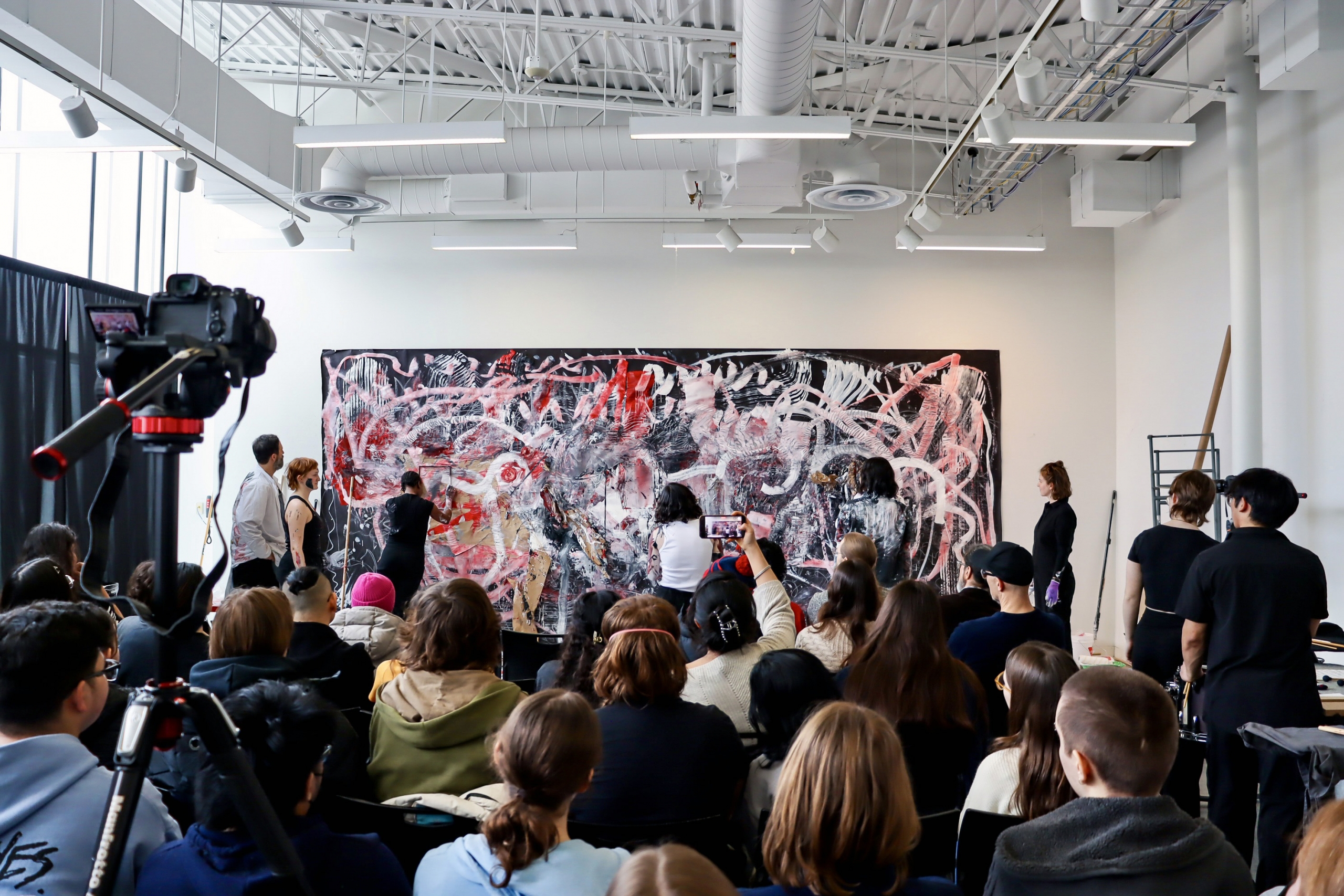 A wide view shows students painting a collaborative artwork during a live performance with an audience present.