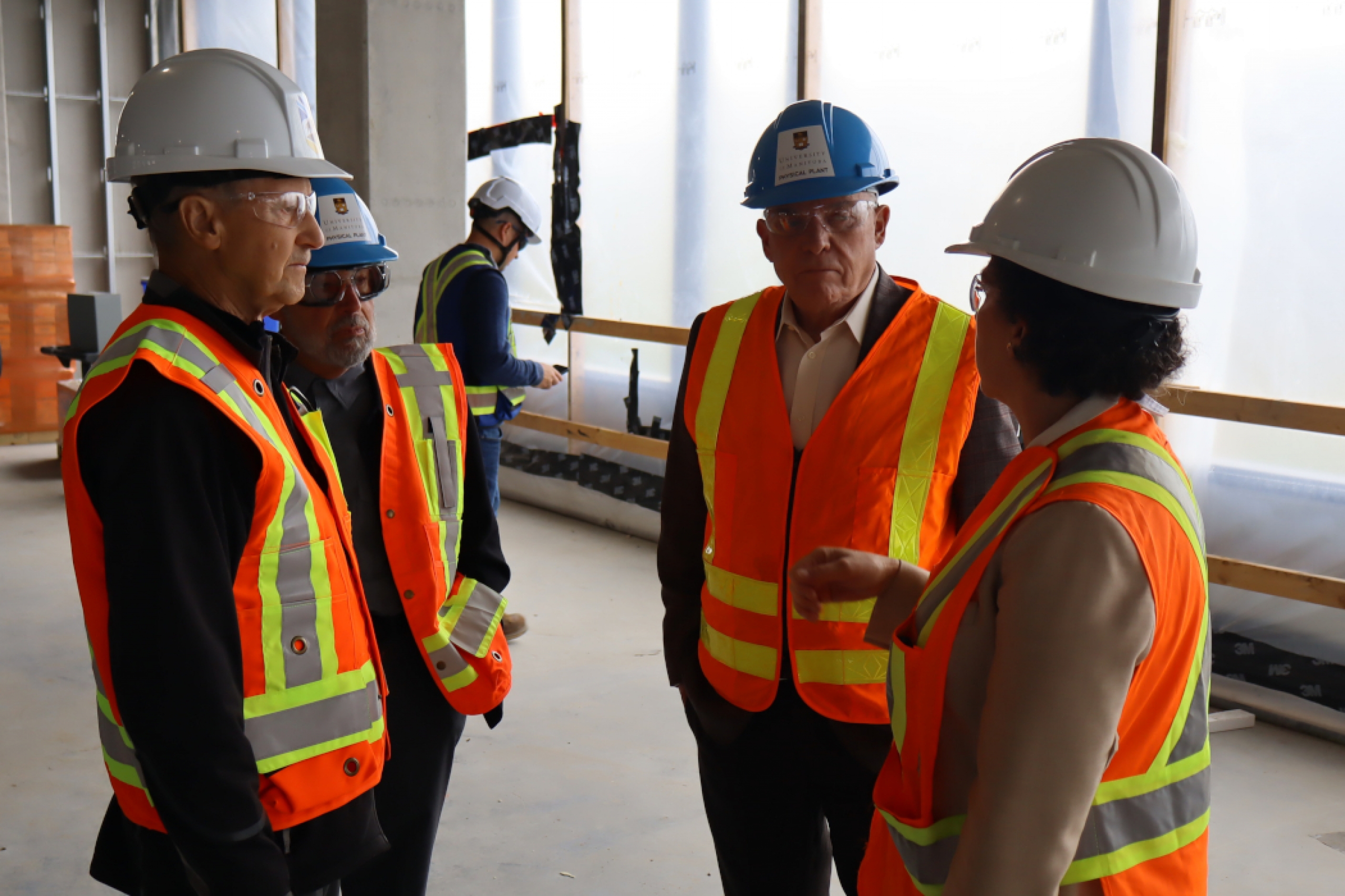 Four people wearing hard hats, safety vests and glasses have a conversation in a building under construction.
