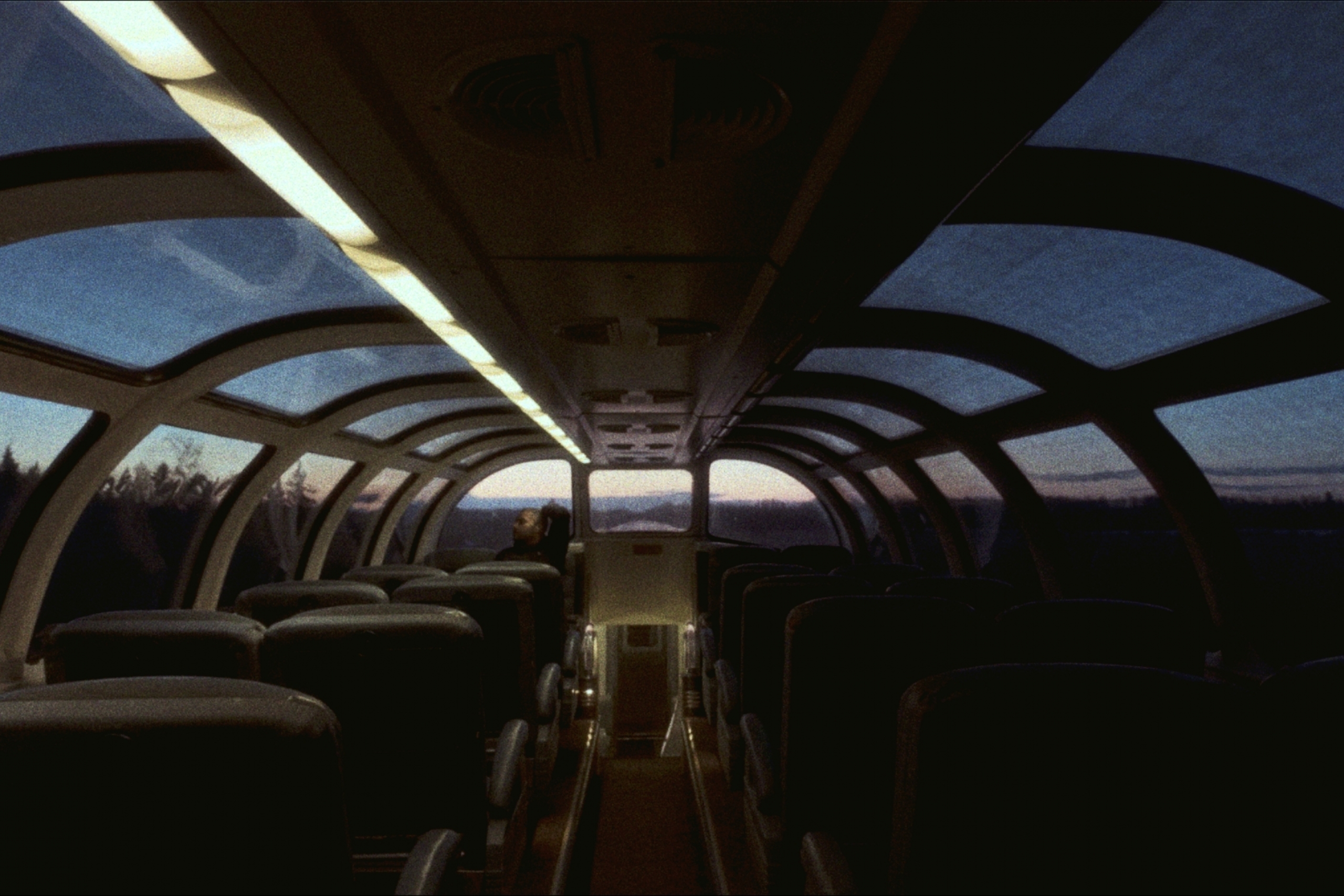 The dim upper cabin of a train as the sun is rising. Only one person sits in the back seat. It is peaceful.
