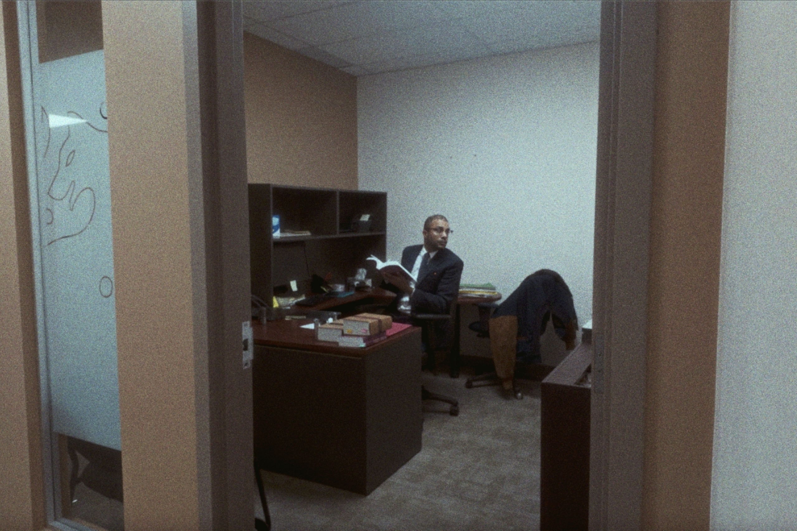 a man in a suit holding a book sits at a desk seen through a beige office door frame.