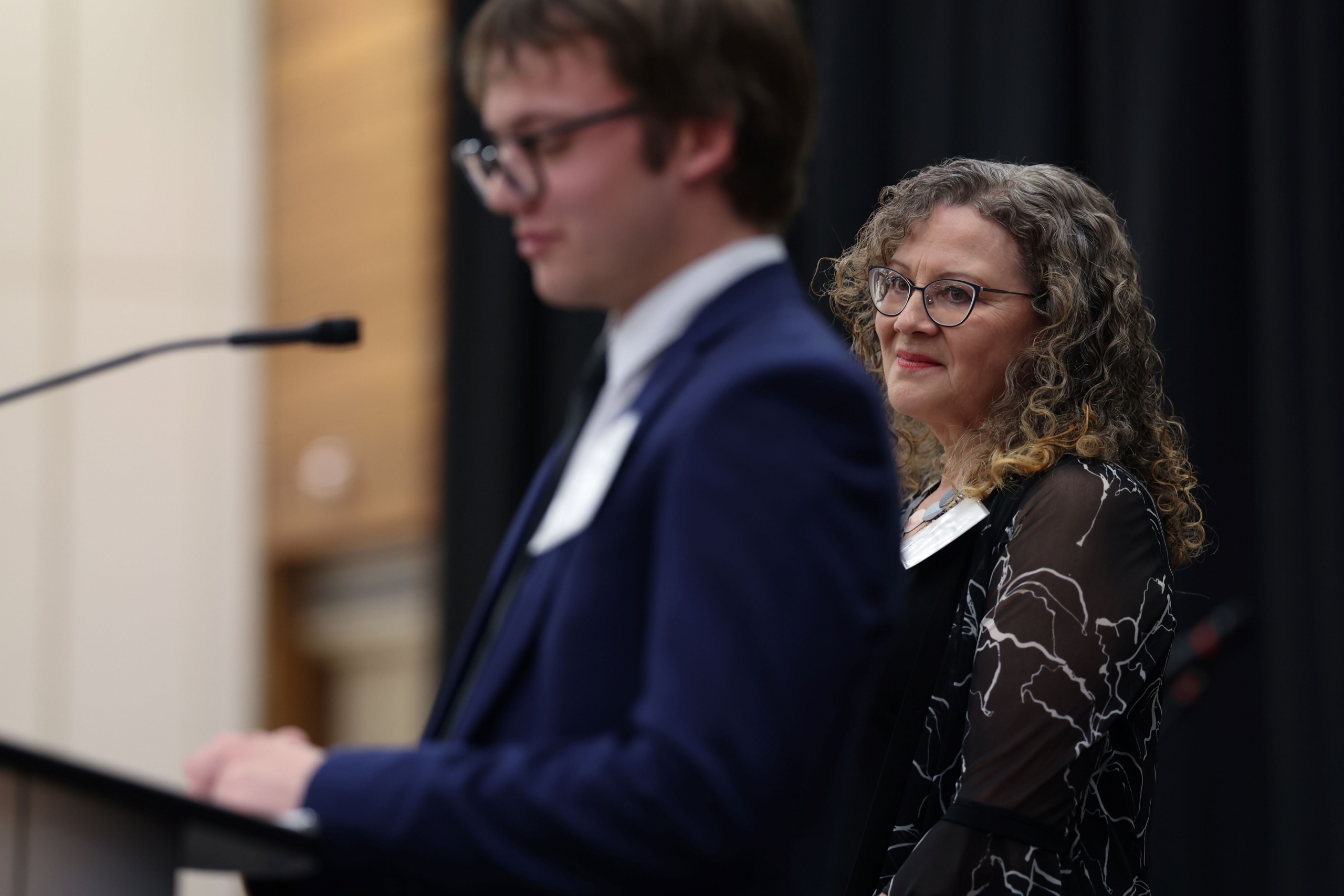 A former educator is looking over the shoulder of her past student while he gives a speech.
