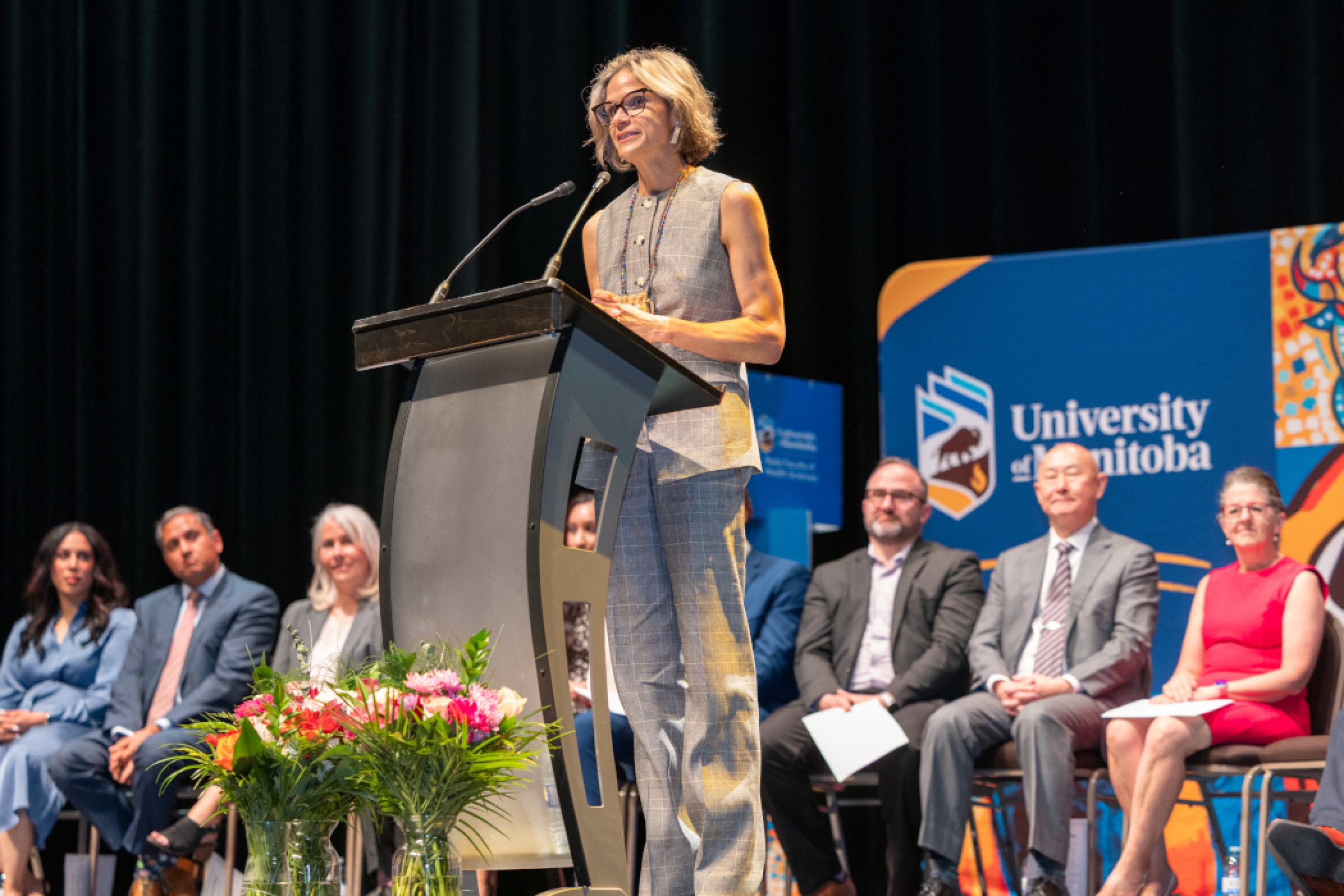 Dr. Sara Goulet speaks at a lectern. Eight health-care professionals sit in chairs behind her. 