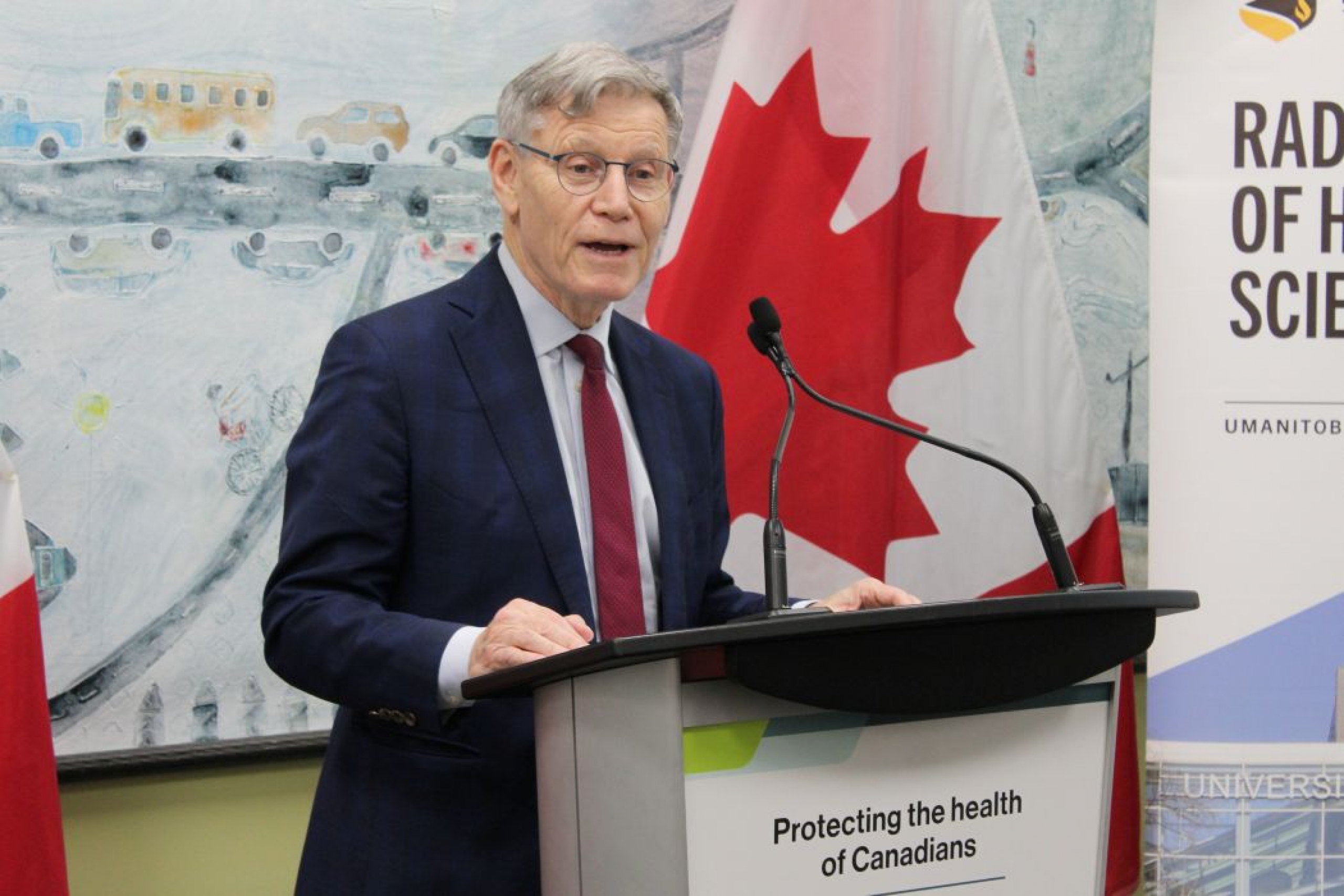 Terry Duguid speaks into two microphones at a lectern. On the front of the lectern is a sign that reads "Protecting the health of Canadians." Behind him are two Canadian flags and a Rady Faculty of Health Sciences banner.