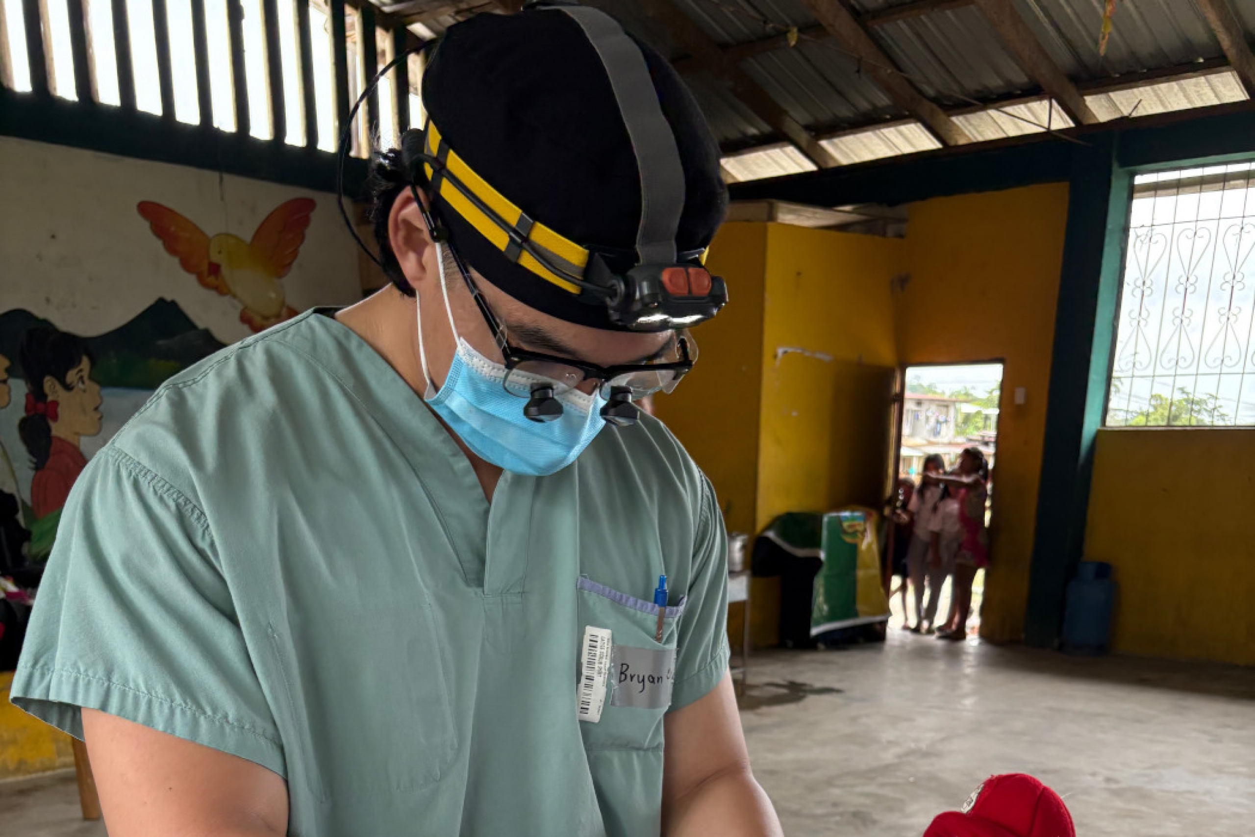 Bryan Kwak treats a young patient. He is wearing a mask, gloves and a headlamp. The child holds a stuffed toy dog.
