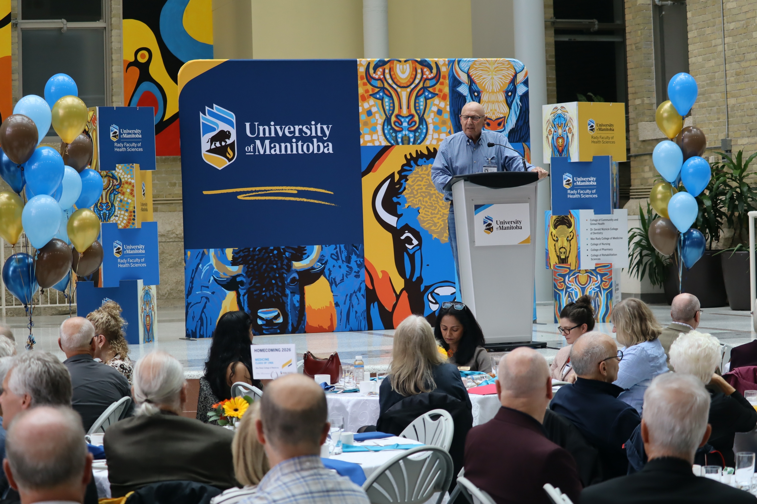 Dr. Brian Postl speaks at a lectern on a stage with a UM banner in the background. 