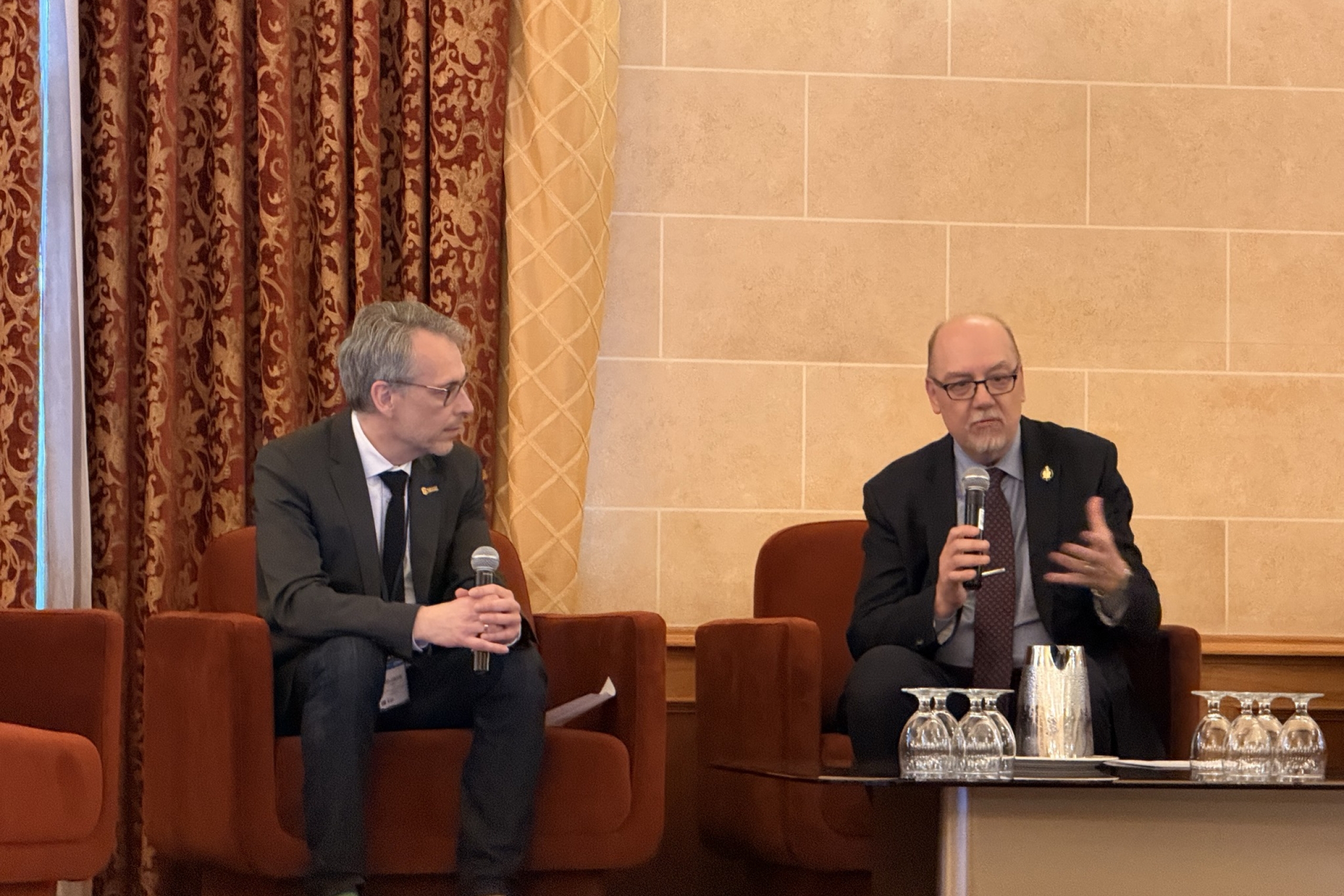 Two speakers seated on stage during a panel discussion, holding microphones in a formal conference setting