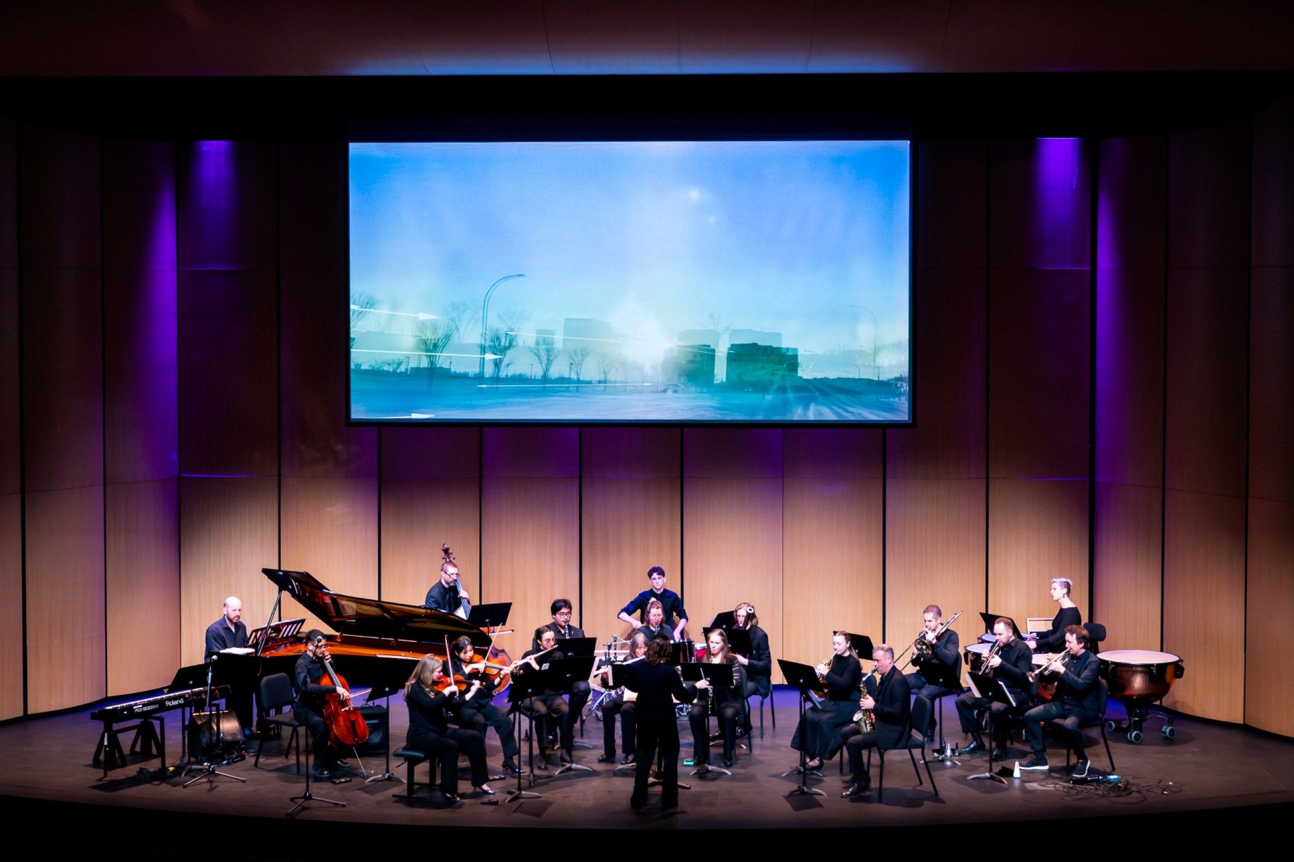 A full ensemble performs on stage as a conductor leads musicians beneath a large projected video image during a ReSound Series concert.