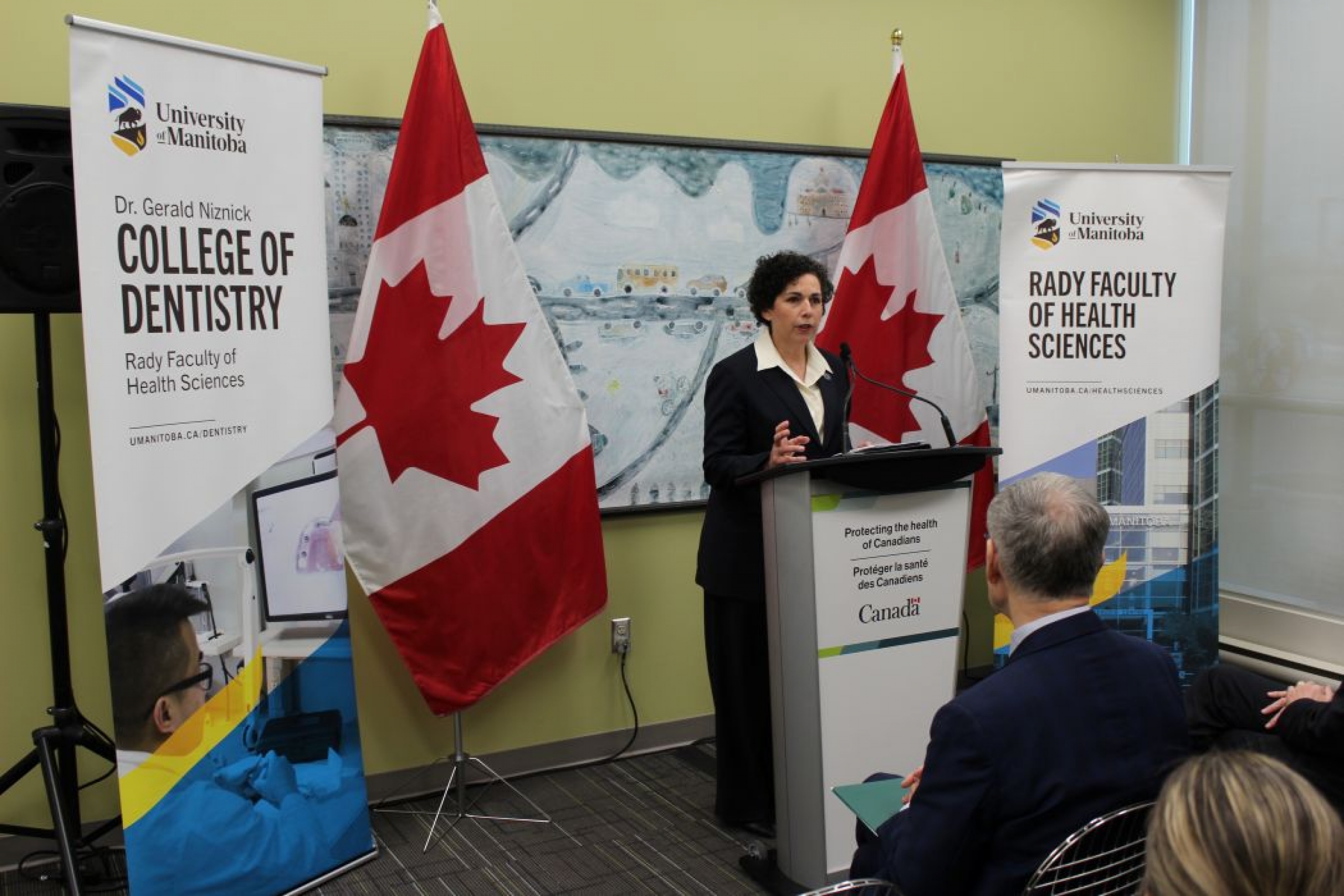 Dr. Anastasia Kelekis-Cholakis speaks into two microphones at a lectern. To her left is a Canadian flag and a Rady Faculty of Health Sciences banner. To her right is a Canadian flag and a Dr. Gerald Niznick College of Dentistry banner.