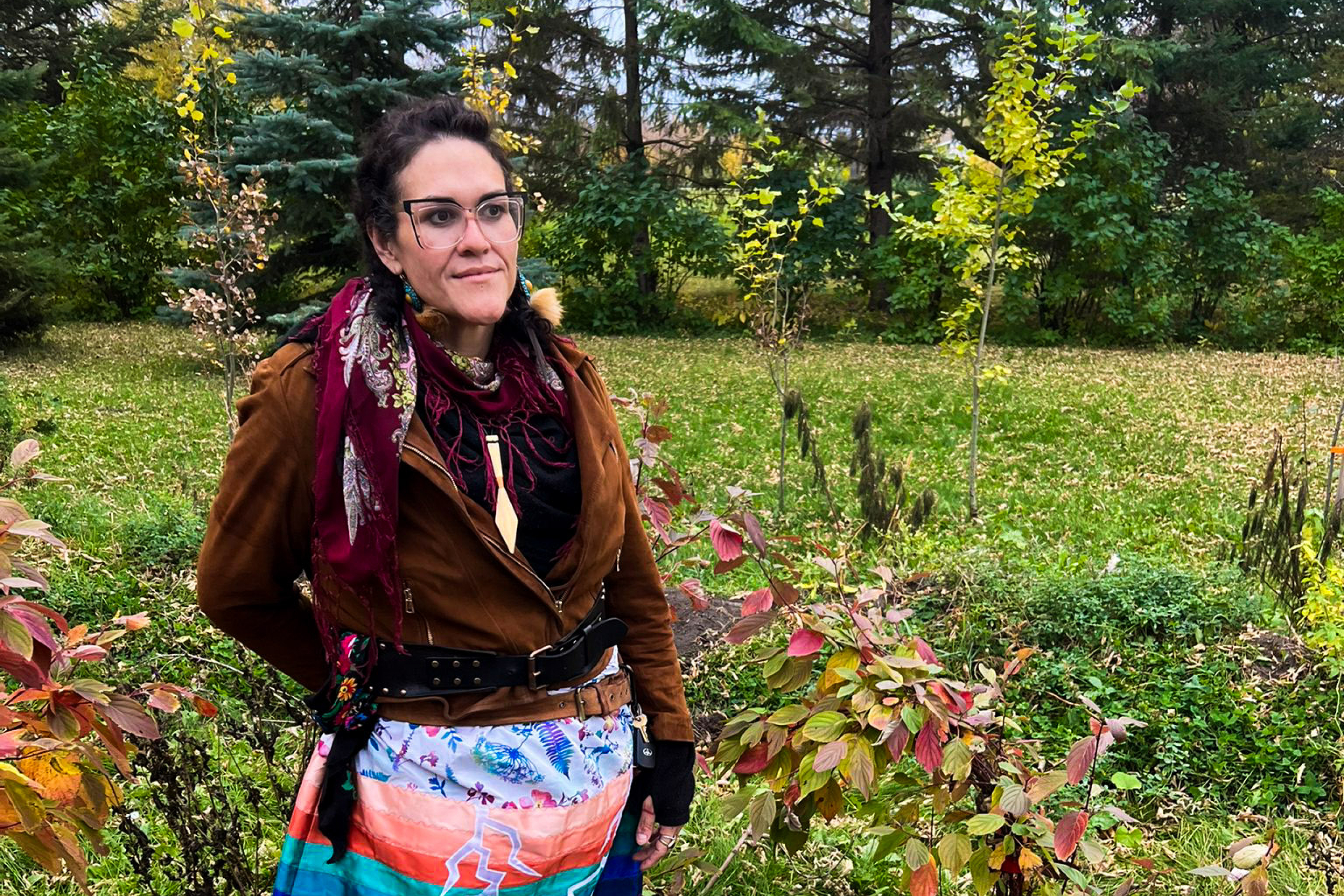Aimee stands outside in nature dressed in a warm jacket and brightly coloured ribbon skirt.