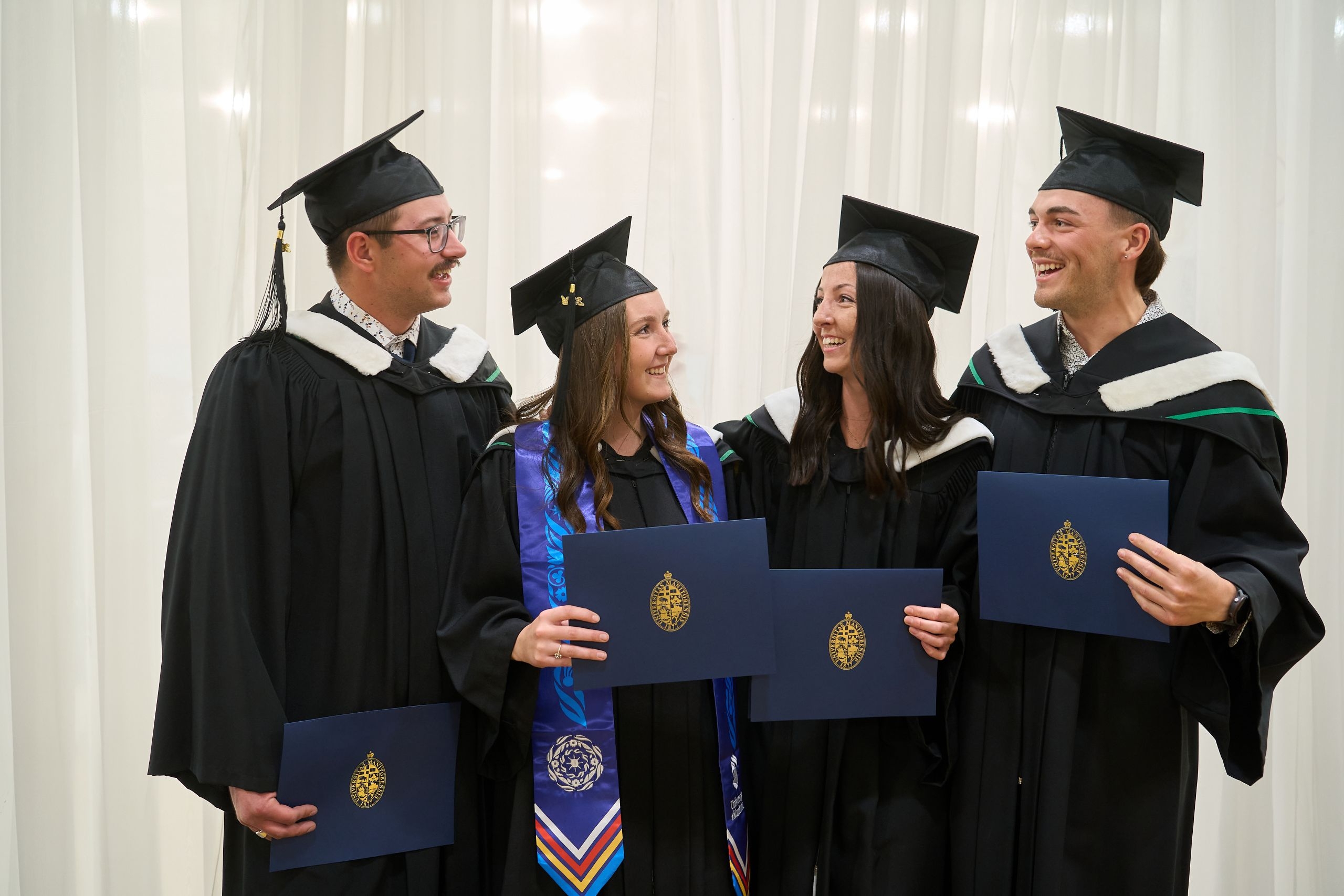 4 graduating students dressed in caps and gowns hold their degrees and celebrate together.