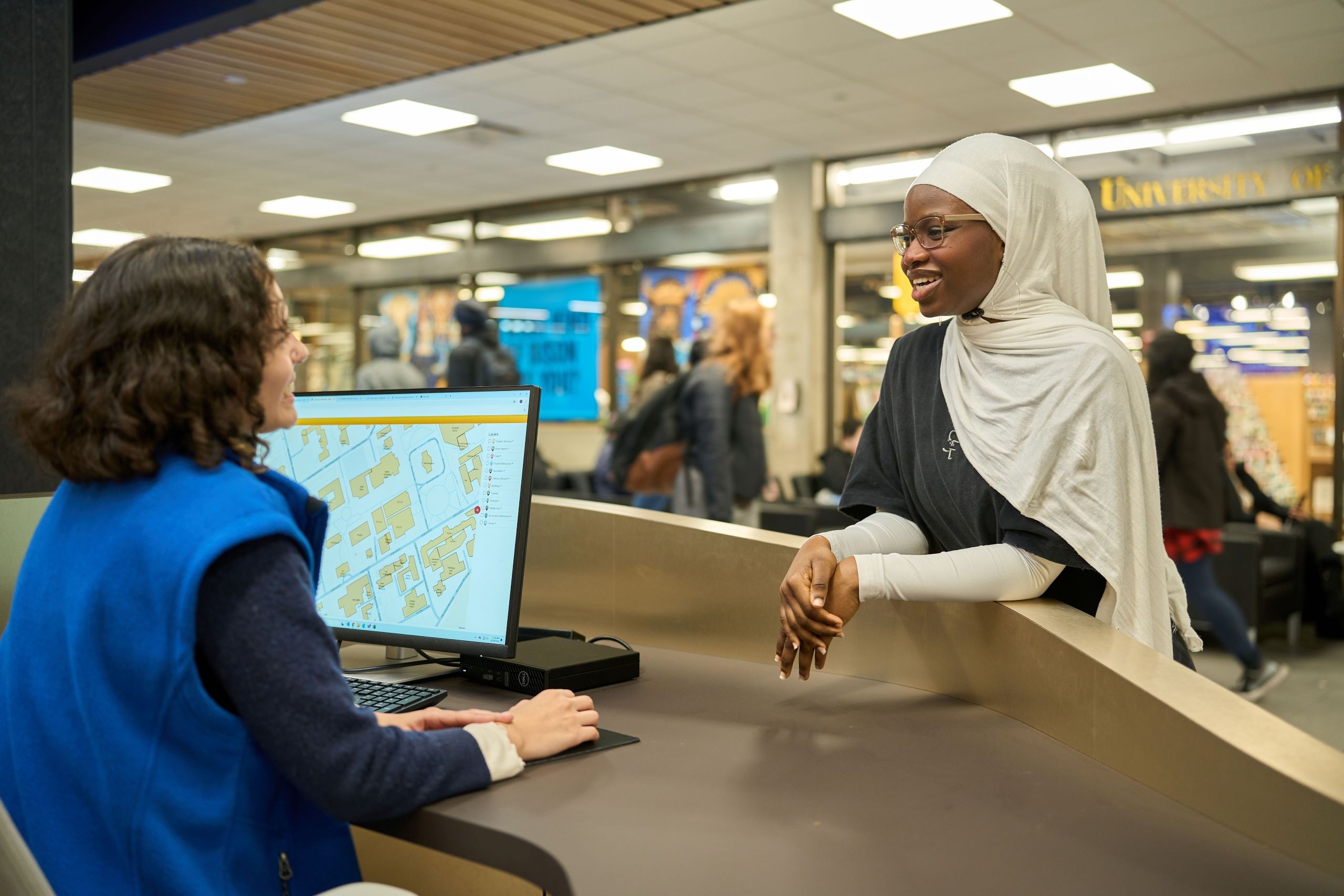 A person wearing a head scarf seeks assistance from a person at an information desk.