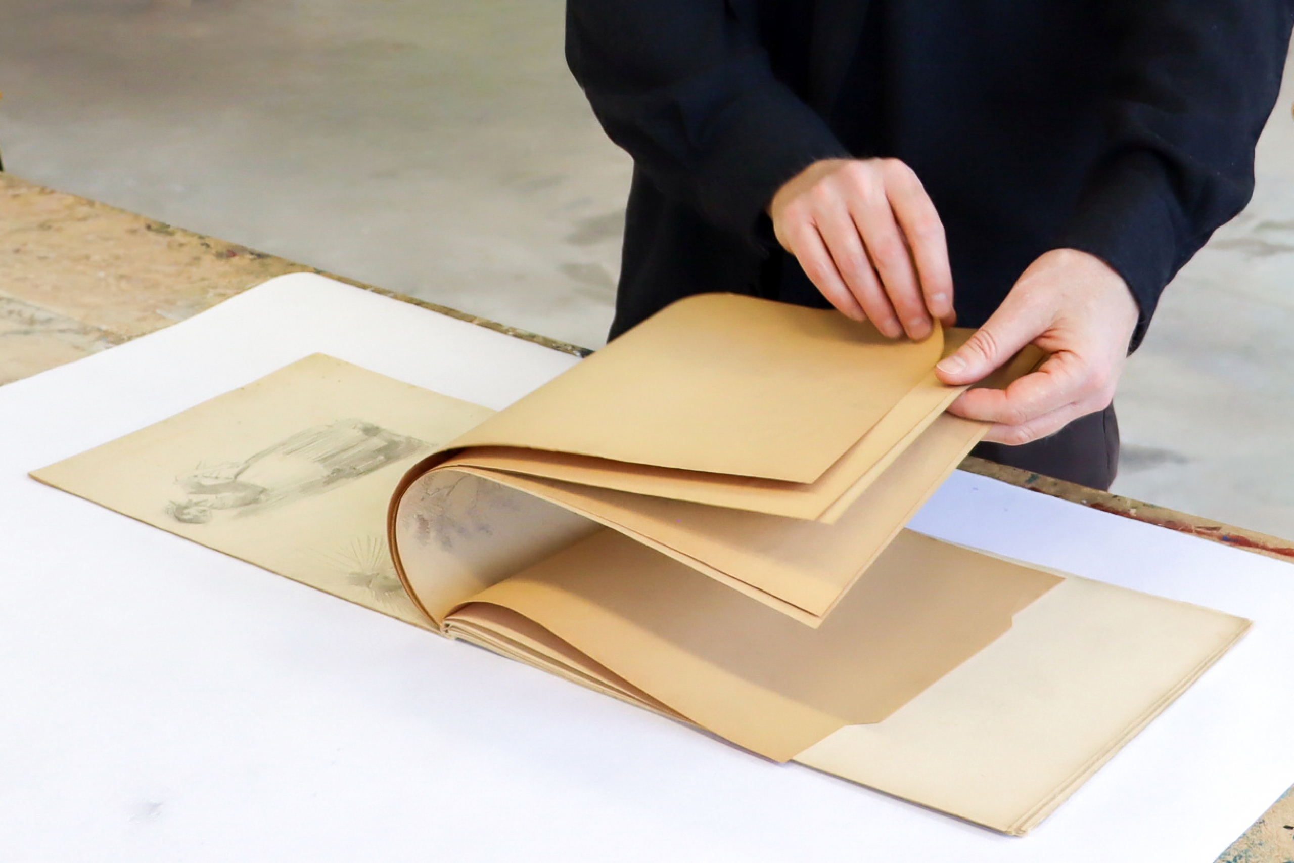 Close-up of Derek Dunlop's hands in a dark jacket turning pages of a large art portfolio on a table, revealing pencil drawings on cream paper.