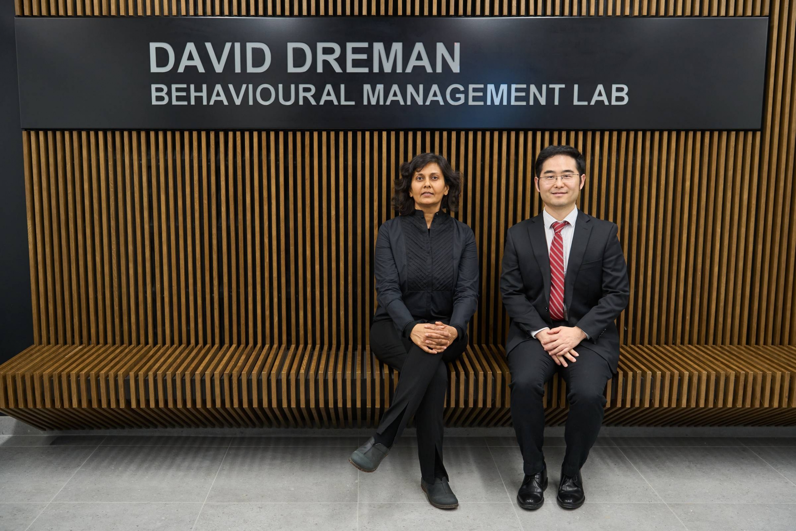 A woman and a man sit in front of the sign for the David Dreman Behavioural Management Lab