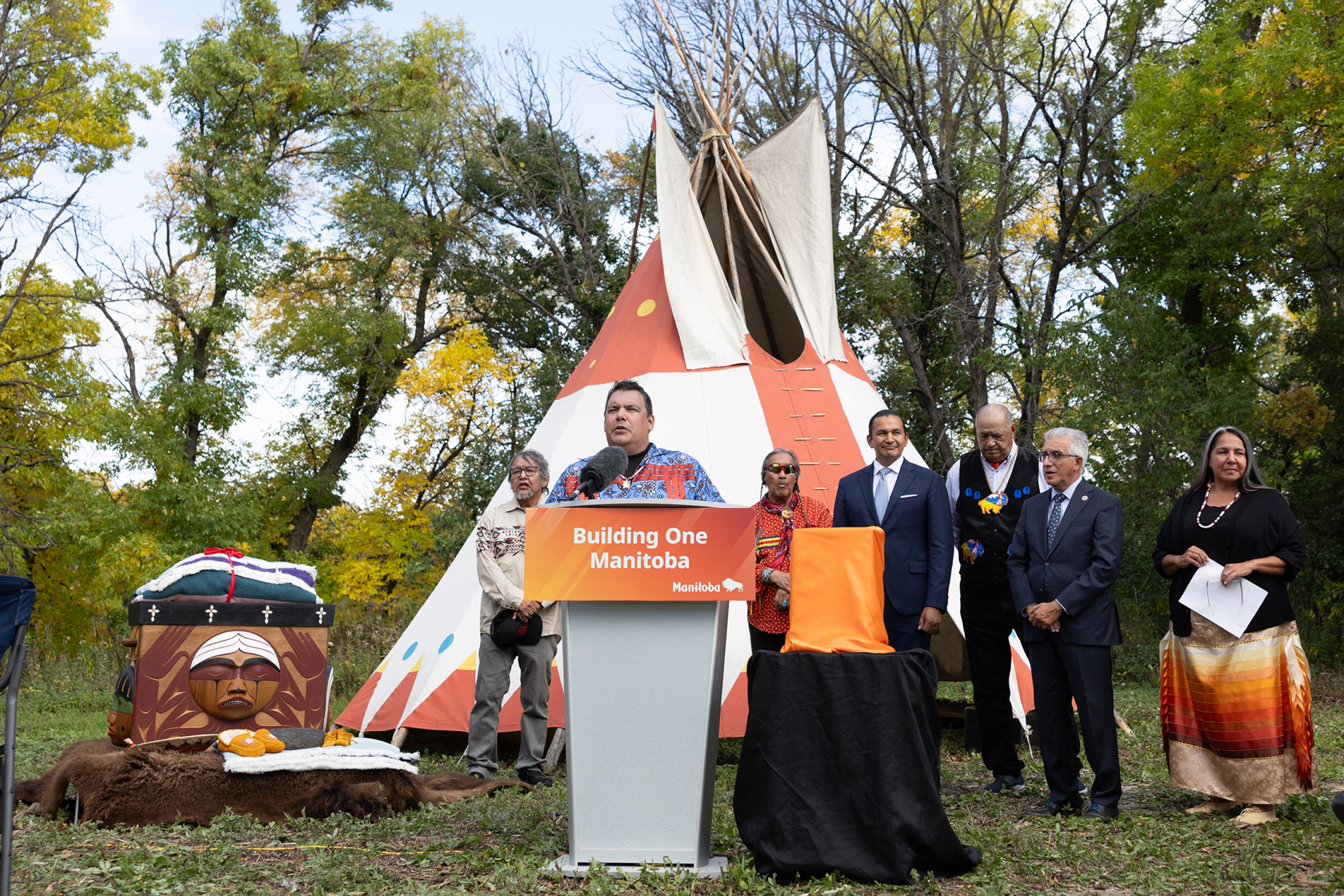 Niigaan Sinclair at podium with Elders, Survivors, UM President Michael Benarroch and Stephanie Scott standing behind him